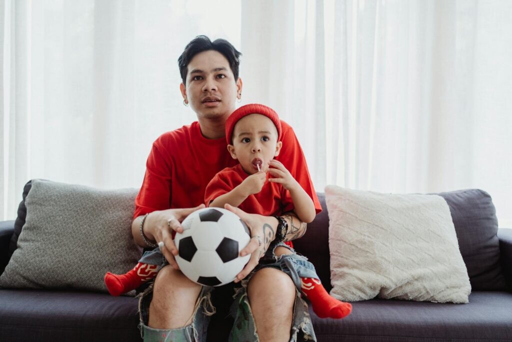 A father and son enjoy quality time together, sharing a lollipop and a soccer ball on the sofa.