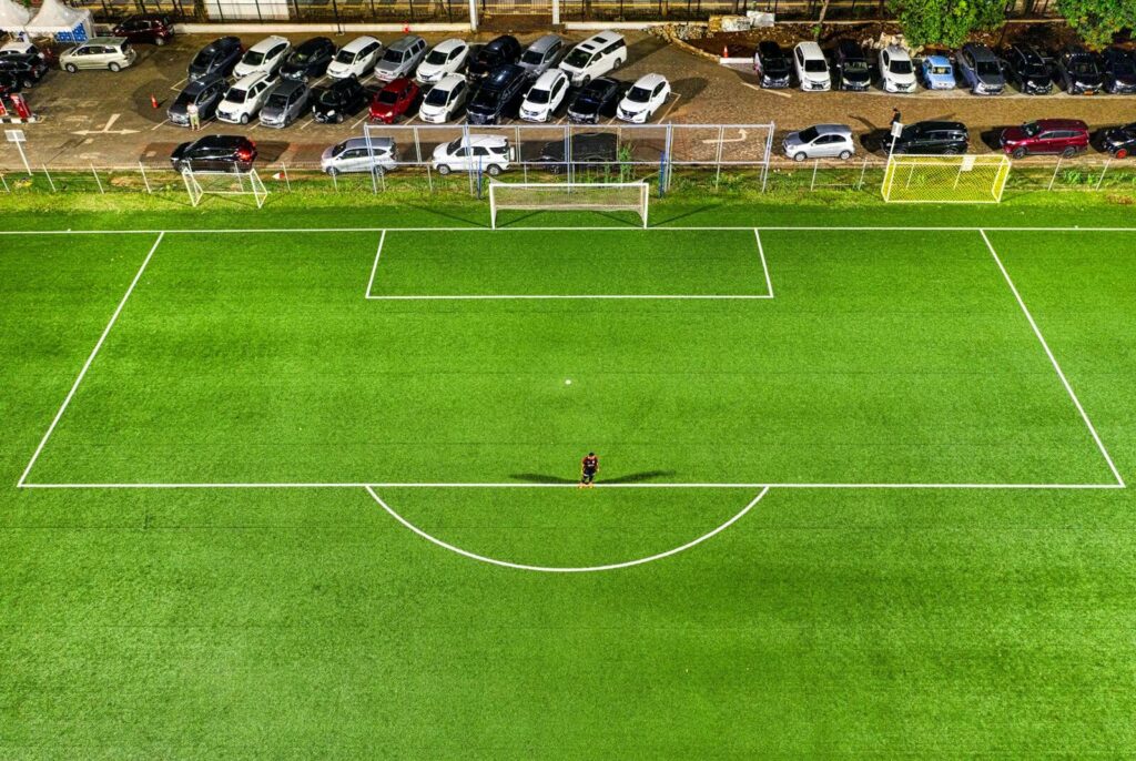 Aerial view of a soccer field at night in Jakarta with a lone player casting a shadow under stadium lights.